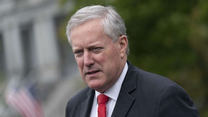 A middle-aged white man in a dark suit, white shirt, and red tie, standing outdoors and speaking to reporters. His expression is serious and focused as he gestures while talking.