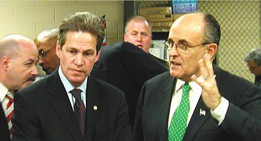 Rudy Giuliani speaks animatedly beside Senator Norm Coleman, surrounded by staff in an indoor setting.