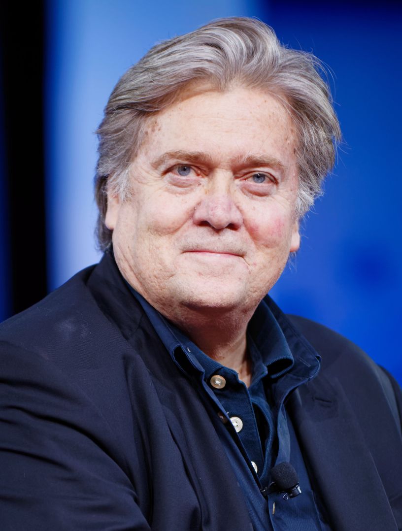 Close-up portrait of Steve Bannon wearing a dark blazer and navy shirt, seated against a blue background at CPAC 2017.
