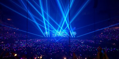 A vast indoor stadium packed with a sea of multicolored wristbands and glow sticks, illuminated by intersecting bright blue laser beams converging on a central stage during a Coldplay concert.