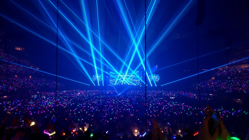 A vast indoor stadium packed with a sea of multicolored wristbands and glow sticks, illuminated by intersecting bright blue laser beams converging on a central stage during a Coldplay concert.