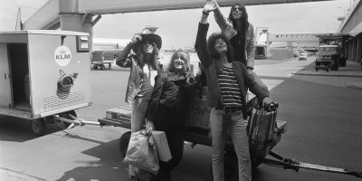 Black-and-white photograph of the four members of Dutch rock band The Golden Earrings standing on an airport luggage cart at Schiphol, waving and smiling toward fans off-camera.