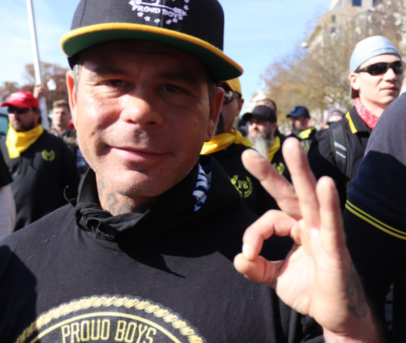 Proud Boys member in branded black and yellow gear making an “OK” hand sign during a 2020 MAGA rally in Washington, D.C., surrounded by other members in similar attire.