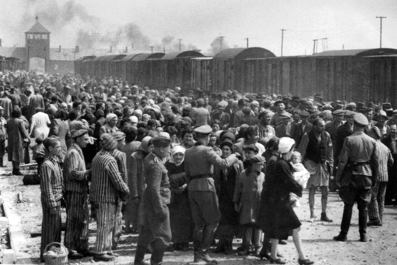 Black-and-white photo of a large crowd of Jewish men, women, and children at Auschwitz-Birkenau in 1944, surrounded by Nazi soldiers conducting a selection. People are being directed to different lines on a dirt ramp, with trains and smoke visible in the background.